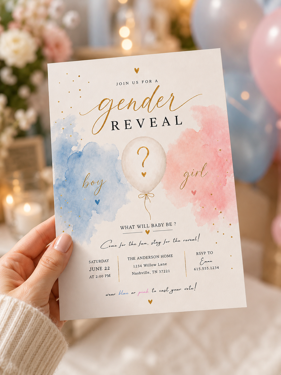 A hand holds a baby shower gender reveal invitation card featuring a watercolor blue and pink design with a white balloon and gold text, set against a softly blurred background of balloons and warm lights.
