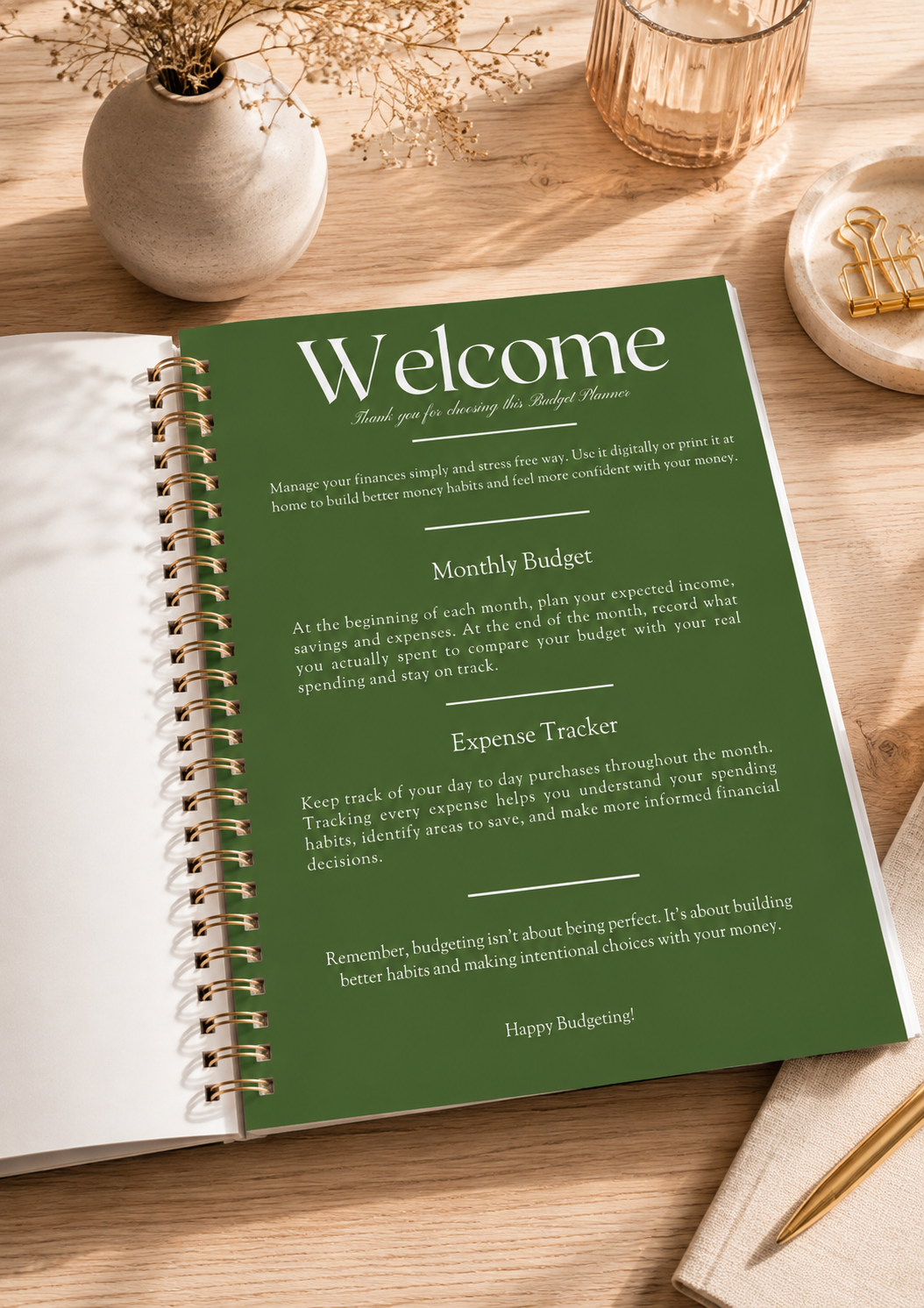 An open spiral-bound planner or notebook on a wooden tabletop, showing a green 'Welcome' page with budgeting and expense tracker text. The scene includes a vase, glass, dish, and pencil as styled props.