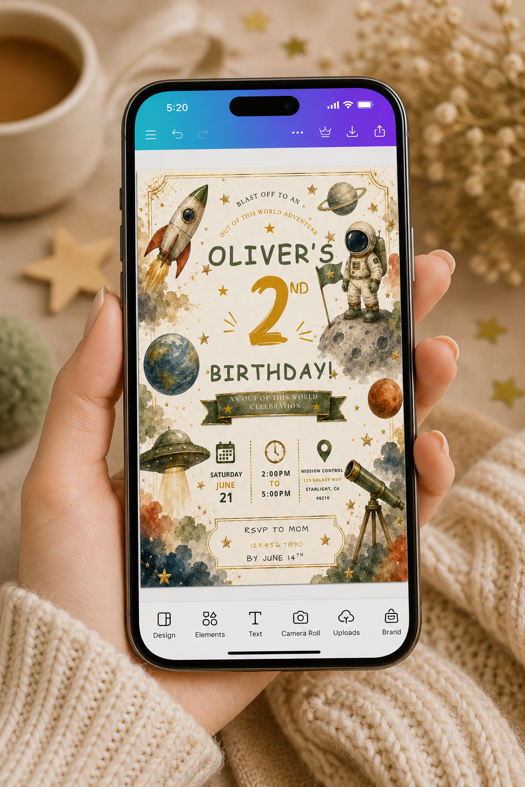A hand holds a smartphone displaying a birthday invitation design with space-themed graphics like rockets, planets, and astronauts. The scene is styled with a cup of coffee, dried flowers, and a beige knit sweater in the background.