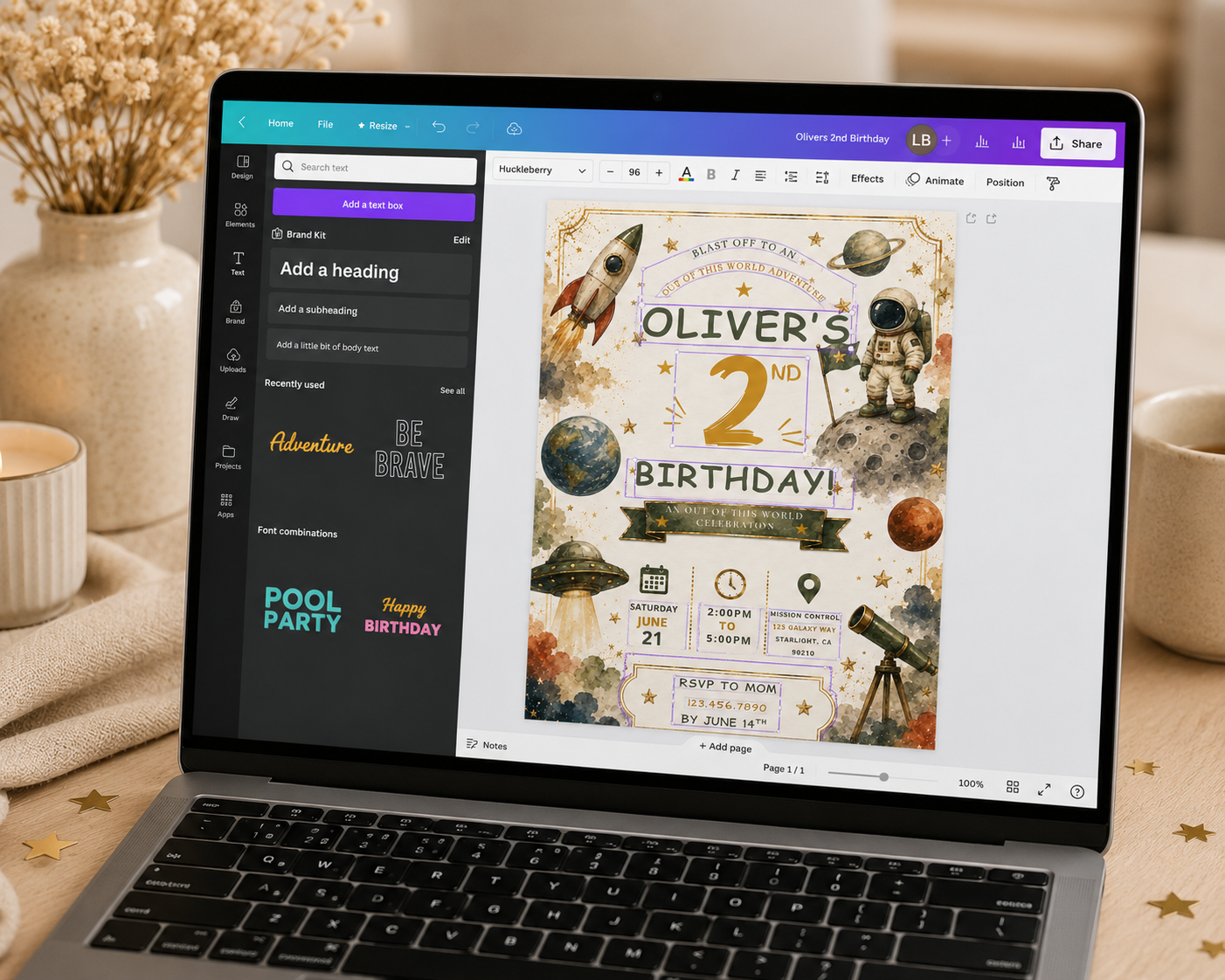 A laptop on a beige tabletop showing a digital birthday invitation design on screen, with a mug, candle, and dried flowers in the softly blurred background.