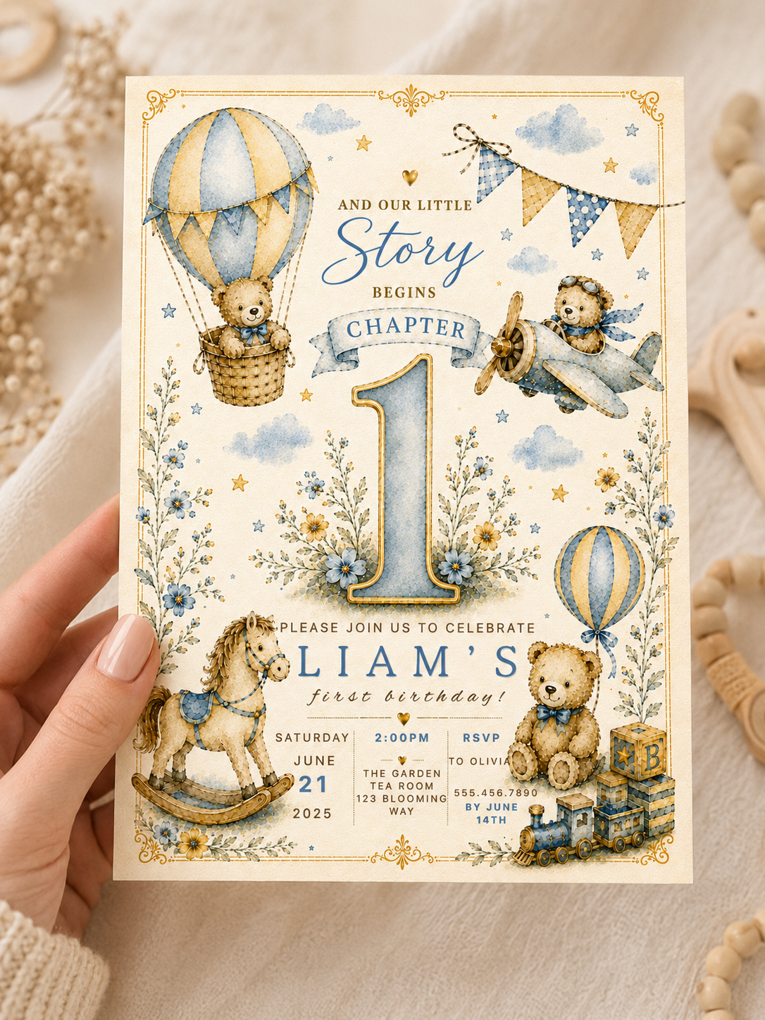 A hand holds a baby first birthday invitation card featuring a vintage-style illustration with teddy bears, hot air balloons, flowers, and a rocking horse on a cream background.