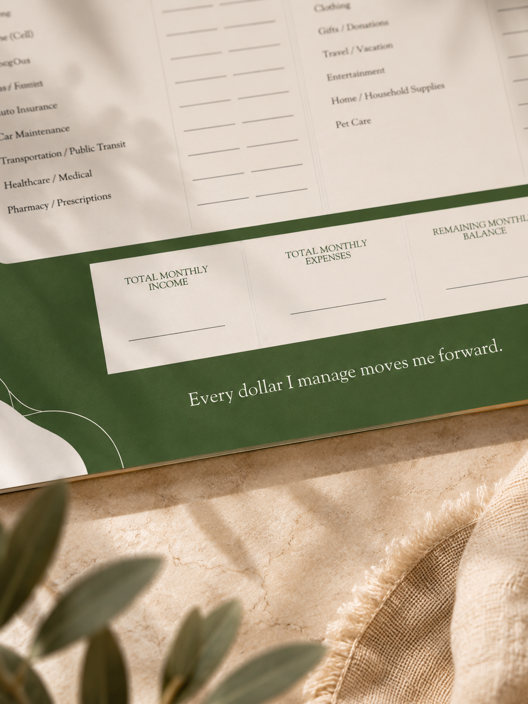 A close-up of a printed budgeting or financial planner page with labeled sections for total monthly income, total monthly expenses, and remaining monthly balance, placed on a beige fabric surface with soft shadows and a green header card.