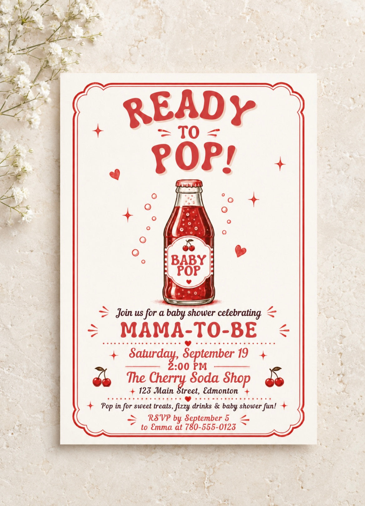 A baby shower invitation card with a vintage red and white 'Ready to Pop!' design featuring a baby soda bottle illustration, styled on a textured light beige surface with small white flowers in the corner.