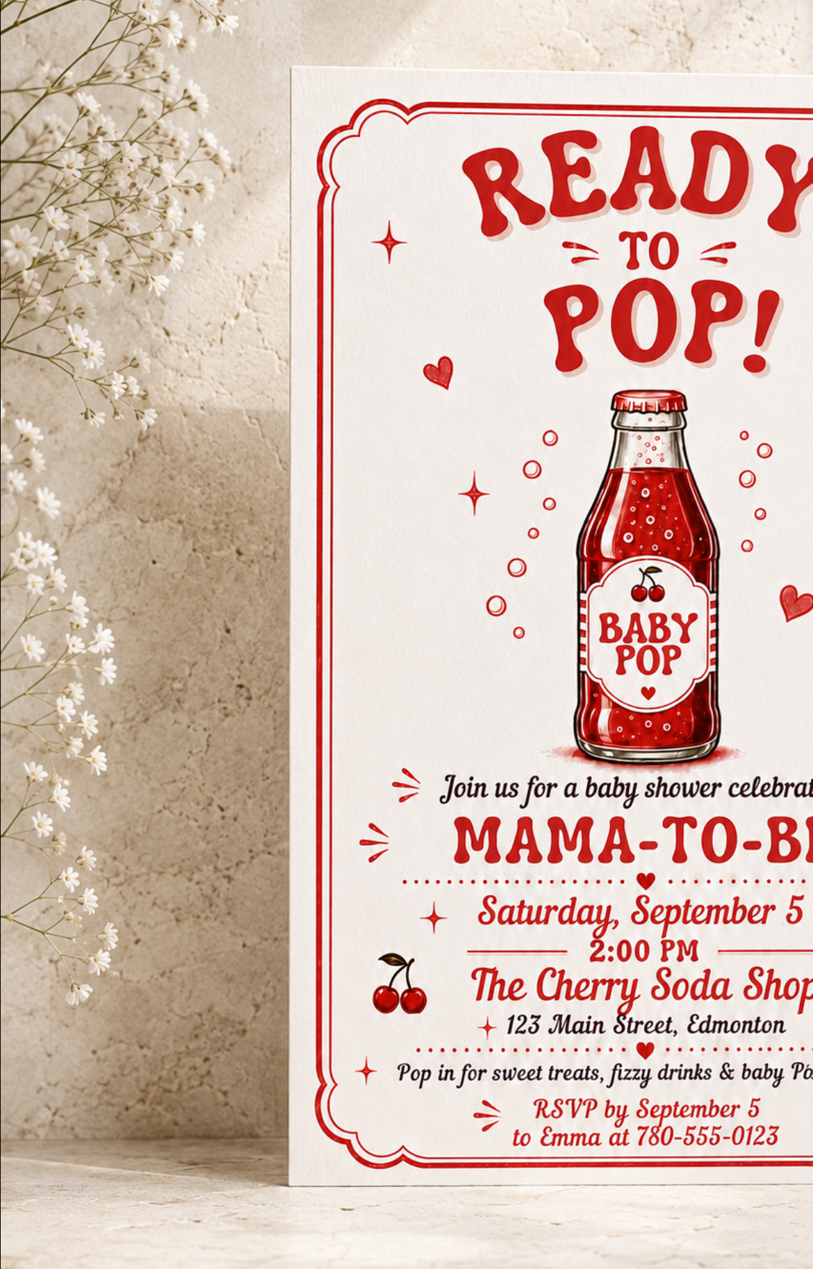 A baby shower invitation card with a red and white design featuring a retro baby pop bottle illustration, styled on a light stone surface with dried baby's breath flowers.