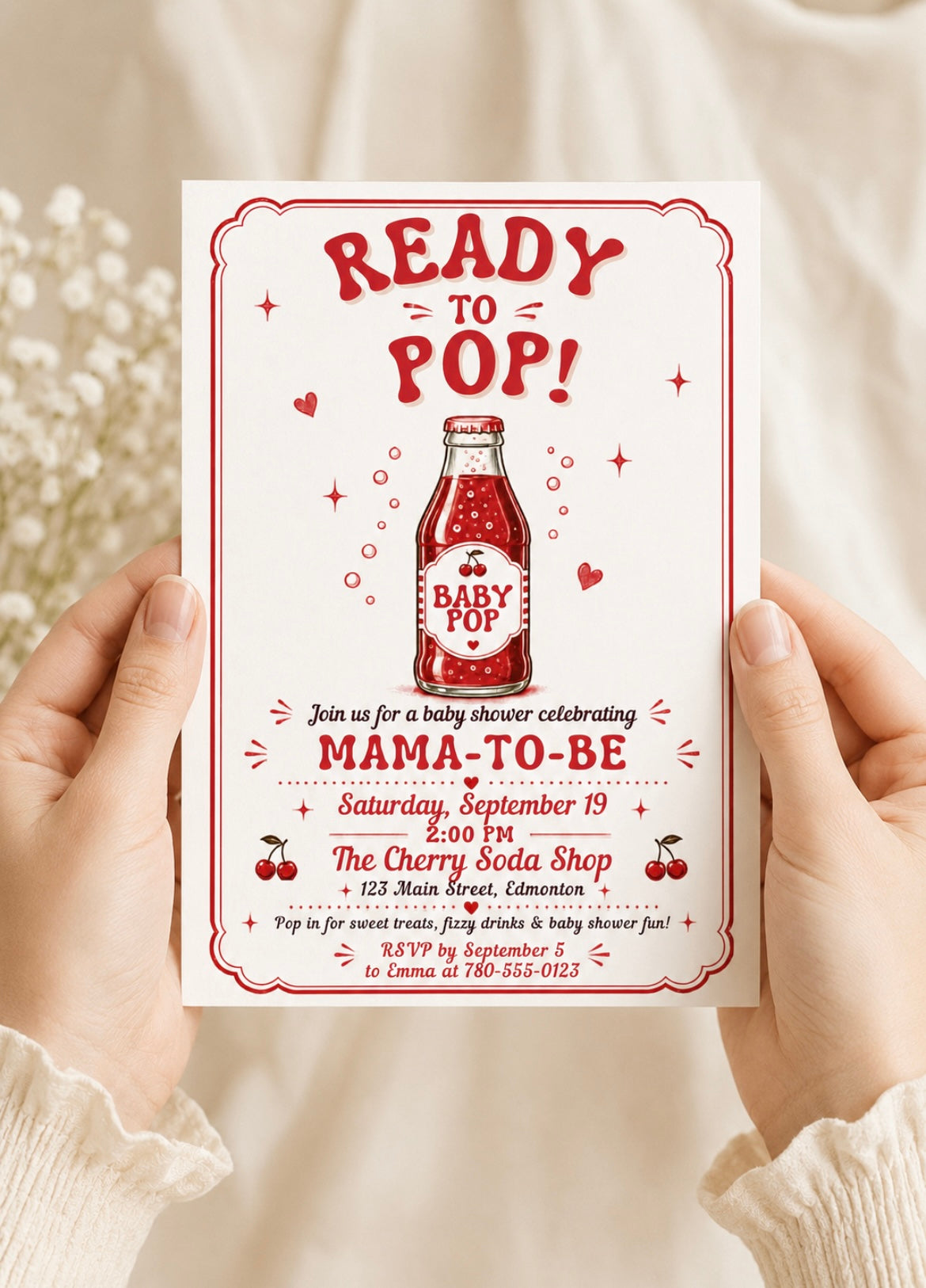 A person holds a baby shower invitation card featuring a retro cherry soda pop design. The card has red text and illustrations on a white background, with the text 'READY TO POP!' and 'MAMA-TO-BE'.
