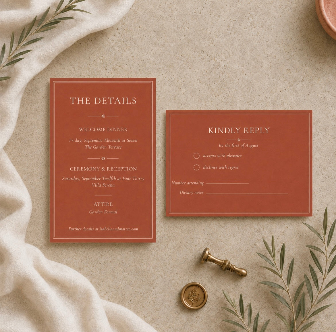 A styled flat-lay of a terracotta red wedding invitation suite on a textured beige surface, surrounded by a cream linen cloth, green leaves, a brass seal stamp, and a copper coin.