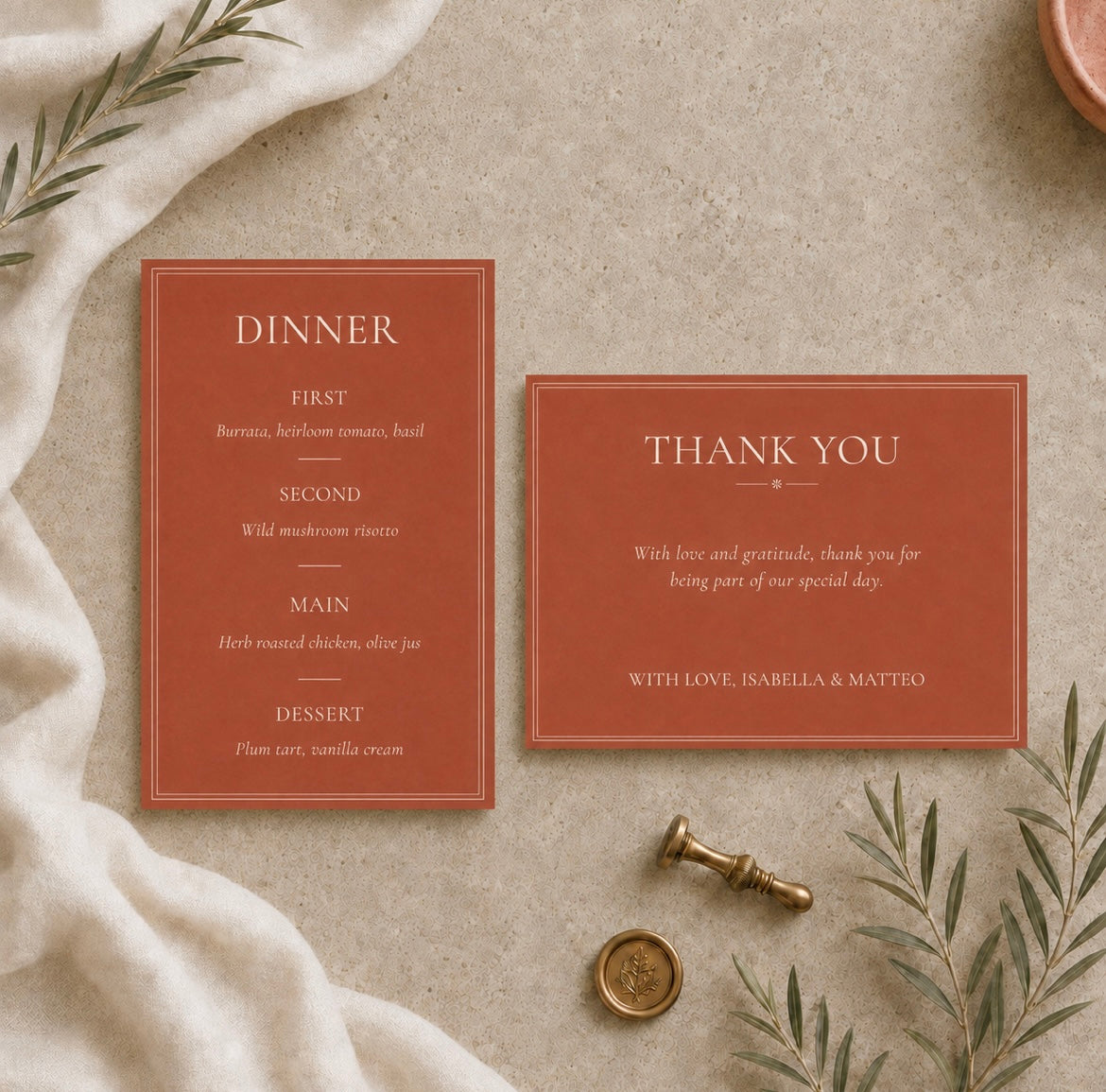 A flat-lay of a matching dinner invitation and thank you card set in a terracotta rust color with elegant cream typography, styled on a textured beige linen surface with botanical sprigs, a wax seal, and a brass stamp.