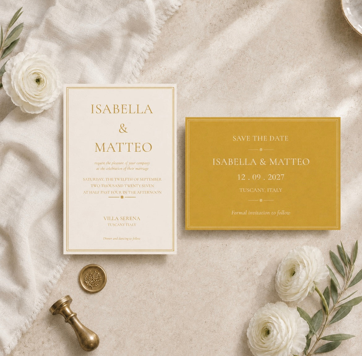 A styled flat-lay of a wedding invitation card and a matching save-the-date card on a textured cream fabric background, surrounded by white flowers, greenery, and a brass stamp.