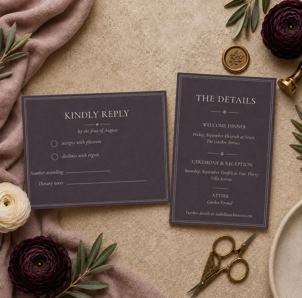 A styled flat-lay of a wedding invitation suite featuring a 'Kindly Reply' card and a 'The Details' card in dark charcoal gray with elegant white typography, surrounded by a blush pink fabric, olive green leaves, deep burgundy flowers, gold scissors, and a brass stamp on a textured beige background.