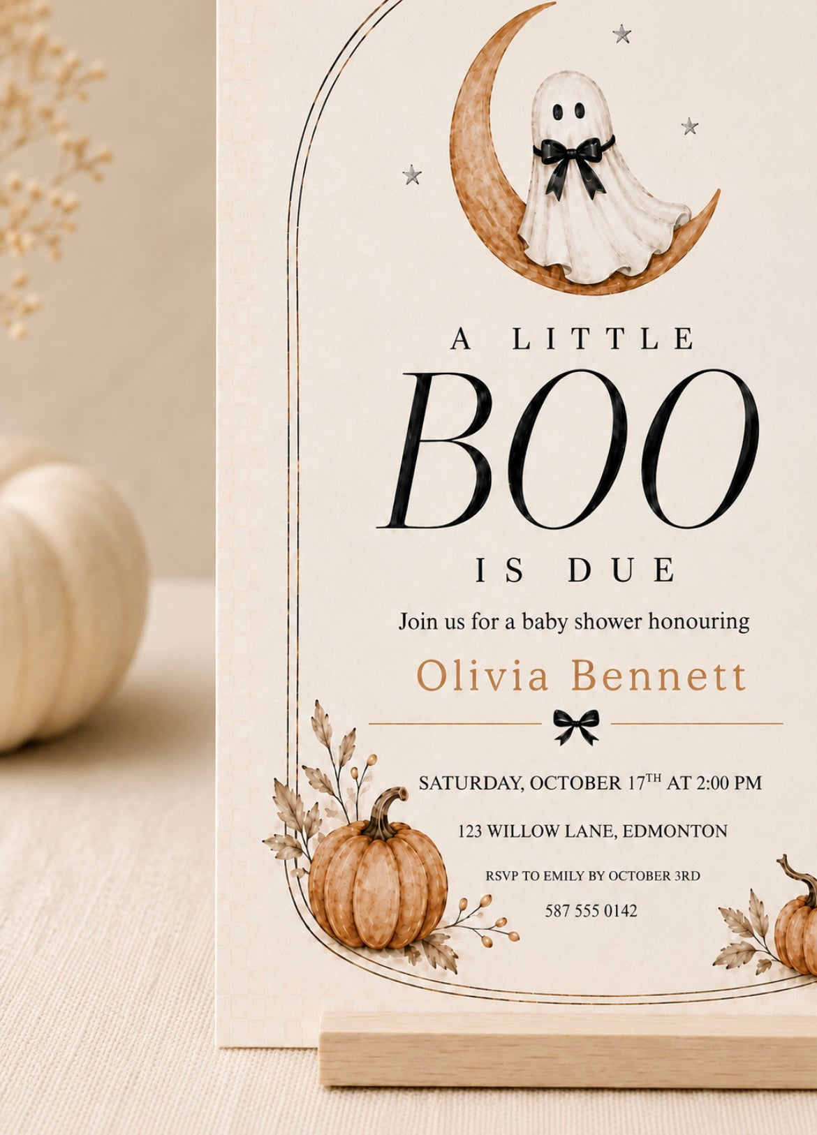 A vertical baby shower invitation card with a Halloween theme, featuring a ghost on a crescent moon, pumpkins, and autumn leaves. The card is styled on a light wooden stand with a neutral beige background and a blurred decorative pumpkin on the left.