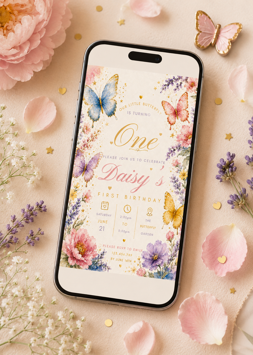 A smartphone lying on a soft pink surface, displaying a colorful first birthday invitation design with butterflies and floral artwork. The phone is surrounded by pink flower petals, lavender sprigs, gold confetti, and decorative butterflies.