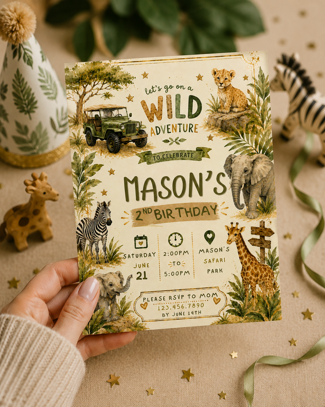 A hand holds a safari-themed birthday invitation card for Mason's 2nd birthday, surrounded by safari props including a zebra toy, a small giraffe figurine, a party hat, and green leaves on a beige fabric background.