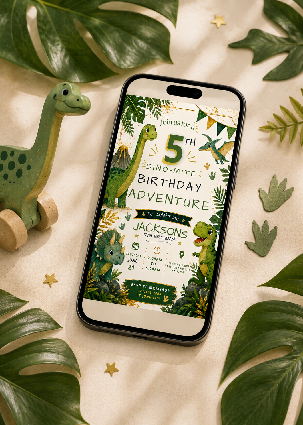 A smartphone lying on a beige fabric surface, displaying a dinosaur-themed birthday invitation design. The scene is styled with large green leaves, small gold stars, a green toy dinosaur, and leaf-shaped props around the phone.