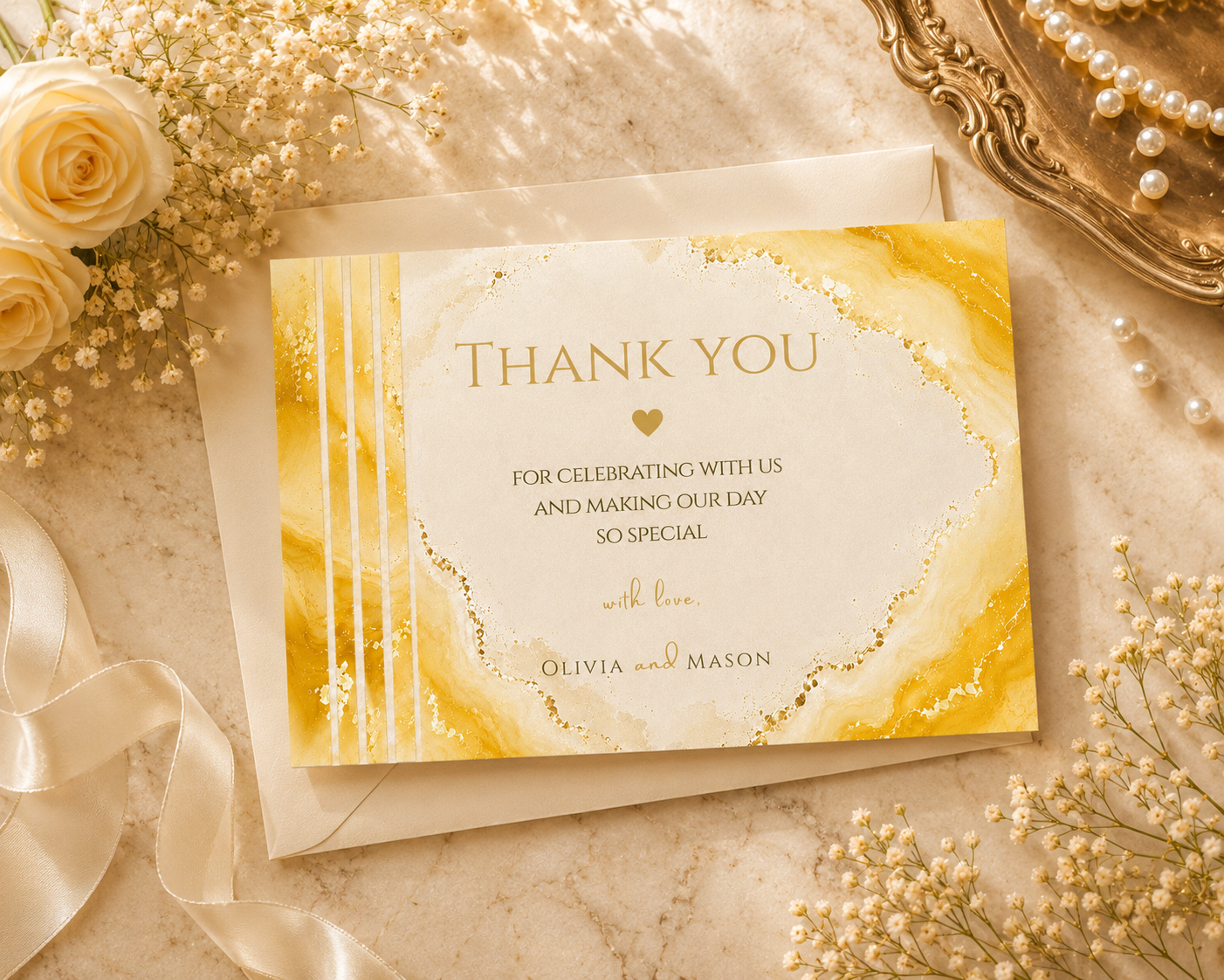 A square thank-you card with a gold and cream marbled design, displayed on top of a matching envelope. The card is styled in a flat lay with white roses, baby's breath, and a gold tray on a marble surface.
